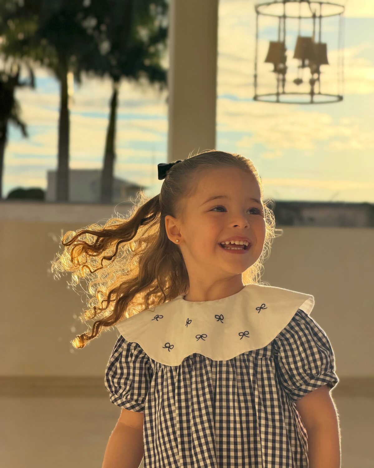 Menina sorrindo com vestido xadrez e gola bordada de laços, ao pôr do sol